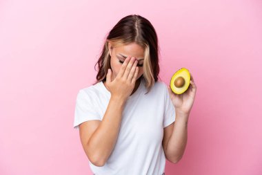 Avokado tutan genç Rus kadın pembe arka planda izole edilmiş, yorgun ve hasta bir ifadeyle.