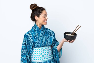 İzole edilmiş beyaz arka planda kimono giyen bir kadın elinde çubuklarla bir kase şehriye tutarken mutlu bir ifade takınıyor.