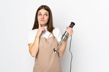 El blenderi kullanan genç bir kadın beyaz arka planda izole edilmiş bir fikir düşünüyor.