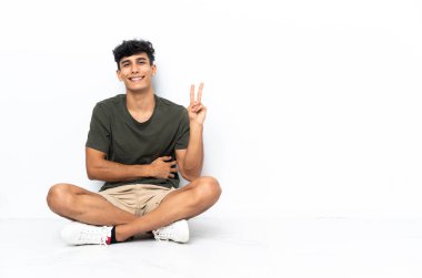 Genç Arjantinli adam yerde oturmuş gülümsüyor ve zafer işareti gösteriyor.