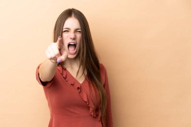 Bej arka planda izole edilmiş genç beyaz kadın hayal kırıklığına uğramış ve ön tarafı işaret ediyor.