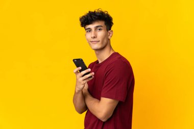 Sarı arka planda izole edilmiş genç Arjantinli bir adam cep telefonu tutuyor ve kollarını kavuşturuyor.