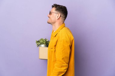 Genç beyaz adam, mor arka planda izole edilmiş şeylerle dolu bir kutuyu kaldırırken yan pozisyonda gülüyor.