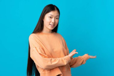 İzole edilmiş mavi arka planda genç bir Çinli kız. Davet etmek için ellerini yana uzatıyor.