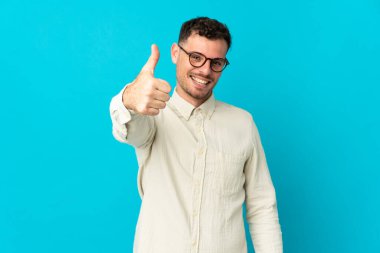 Beyaz tenli yakışıklı adam mavi arka planda izole edildi. Başparmakları havada, çünkü iyi bir şey oldu.