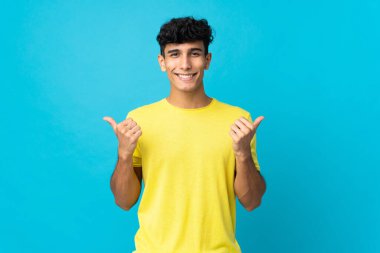 Arjantinli genç adam arka planda izole edilmiş. Baş parmakları havada ve gülümsüyor.