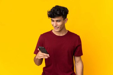 Arjantinli genç adam sarı arka planda izole edilmiş cep telefonuyla mesaj ya da e-posta gönderiyor.