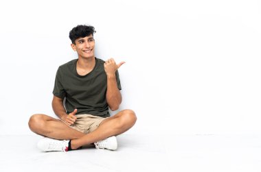 Yerde oturan genç Arjantinli bir adam bir ürünü sunmak için kenarı işaret ediyor.