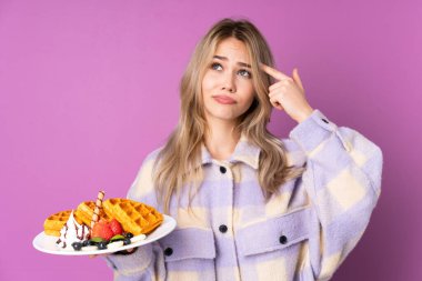 Mor arka planda izole edilmiş waffle tutan genç Rus kız deliliğin jestini yapıyor. Parmağını kafaya koyuyor.
