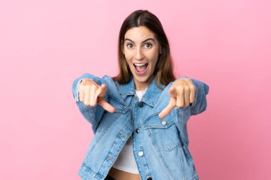 Pembe arka planda izole edilmiş genç beyaz kadın şaşırmış ve ön tarafı işaret ediyor.