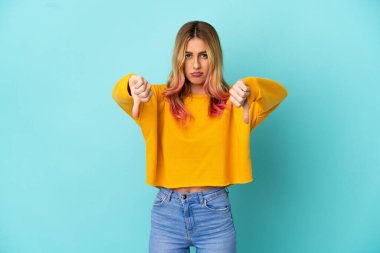 İzole edilmiş mavi arka planda genç bir kadın baş parmağını iki eliyle aşağı gösteriyor.