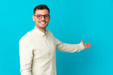 Beyaz tenli yakışıklı adam mavi arka planda izole edilmiş. Davet etmek için ellerini yana uzatıyor.