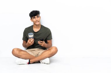 Genç Arjantinli bir adam elinde kahve ve bir şeyler düşünürken cep telefonu ile yerde oturuyor.