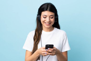 Genç esmer kız izole edilmiş mavi arka planda müzik dinliyor ve cep telefonu arıyor.
