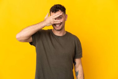 Beyaz tenli yakışıklı adam sarı arka planda izole edilmiş gözlerini elleriyle kapatmış ve gülümsüyor.