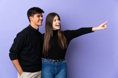 İzole edilmiş mor arka planda genç bir çift parmak göstererek bir ürün sunuyor.
