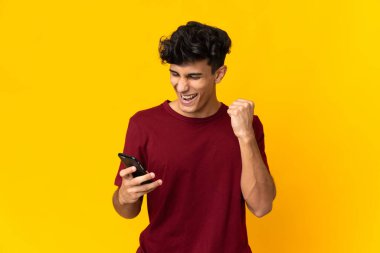 Arjantinli genç adam sarı arka planda cep telefonu kullanıyor ve zafer jesti yapıyor.