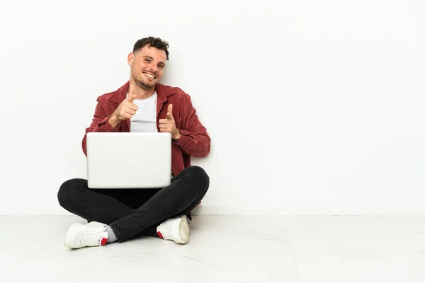 年轻英俊的高加索男子坐在地板上 笔记本电脑指向前面 面带微笑 — 图库照片