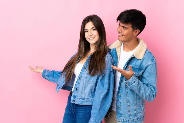 İzole edilmiş pembe arka planda genç bir çift geriye doğru işaret ediyor ve bir ürün sunuyorlar