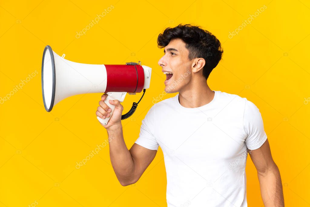 Young Argentinian man isolated on yellow background shouting through a megaphone to announce something in lateral position