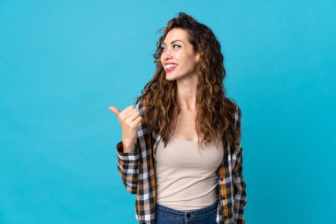 Mavi arka planda izole edilmiş genç beyaz kadın bir ürünü sunmak için kenarı işaret ediyor.