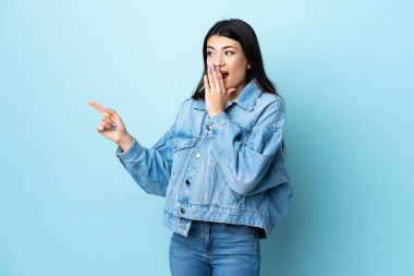 Soyutlanmış mavi arka planda genç esmer kız. Sürpriz bir ifadeyle işaret ederken...