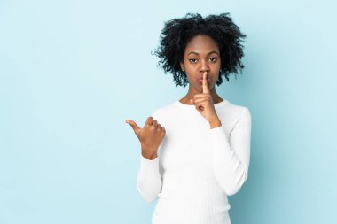 Genç Afrikalı Amerikalı bir kadın mavi arka planda izole edilmiş. Yan tarafı işaret ediyor ve sessizlik hareketi yapıyor.