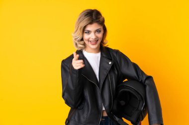 Sarı arka planda izole edilmiş motosiklet kaskı tutan genç bir kadın sana parmak atıyor.