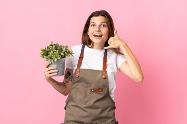 Bahçıvan kadın pembe arka planda izole edilmiş bir bitki tutuyor ve telefon jesti yapıyor. Bana geri dönüş işareti ver.