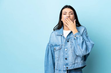 Genç esmer kız izole edilmiş mavi arka planda esniyor ve geniş ağzı eliyle kapatıyor.