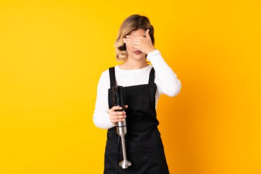 El blenderi kullanan kız sarı arka planda izole edilmiş, gözleri elle kapatıyor. Bir şey görmek istemiyorum.