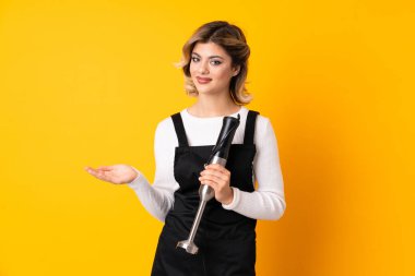 El blenderi kullanan kız sarı arka planda izole edilmiş, davet edilmek için ellerini yana uzatıyor.