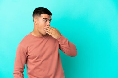 Soyutlanmış mavi arka planda genç yakışıklı bir adam yan tarafa bakarken sürpriz jest yapıyor.