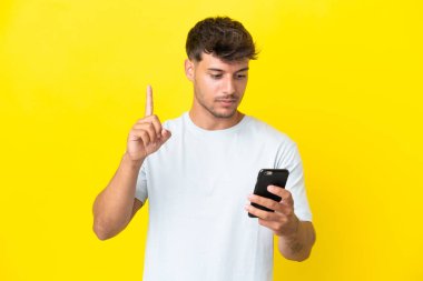 Sarı arka planda izole edilmiş genç beyaz adam cep telefonu kullanıyor ve parmağını kaldırıyor.