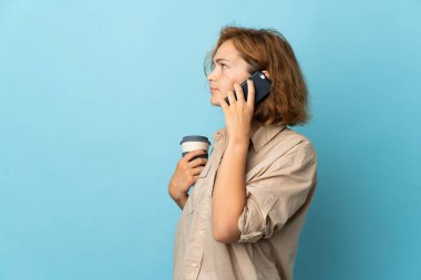 Genç Gürcü bir kadın mavi arka planda izole edilmiş. Elinde kahve ve cep telefonu var.