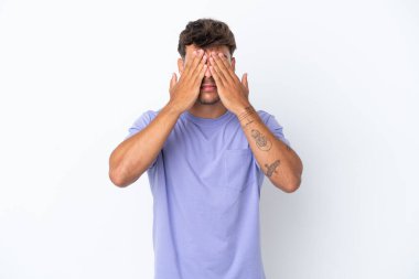 Beyaz arka planda izole edilmiş beyaz tenli yakışıklı adam gözlerini elleriyle kapatıyor.
