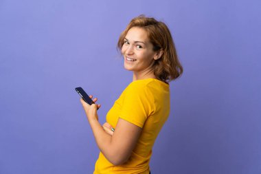 Mor arka planda izole edilmiş genç Gürcü bir kadın cep telefonu tutuyor ve kollarını kavuşturuyor.