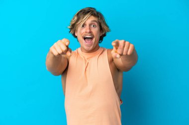 Yakışıklı sarışın adam mavi arka planda izole edilmiş, şaşırmış ve ön tarafı işaret ediyor.