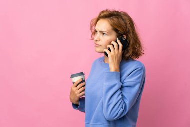 Genç Gürcü bir kadın pembe arka planda izole edilmiş. Elinde kahve ve cep telefonu var.