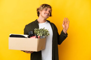 Sarışın adam, sarı arka planda izole edilmiş şeylerle dolu bir kutuyu kaldırırken hareket ediyor. Mutlu bir ifadeyle selam veriyor.