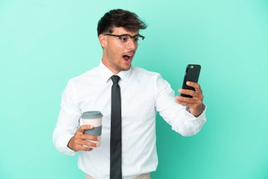 Kafkasyalı iş adamı mavi arka planda izole edilmiş. Elinde kahve ve cep telefonu var.