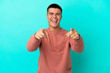 Soyutlanmış mavi arka planda genç yakışıklı adam şaşırmış ve ön tarafı işaret ediyor.