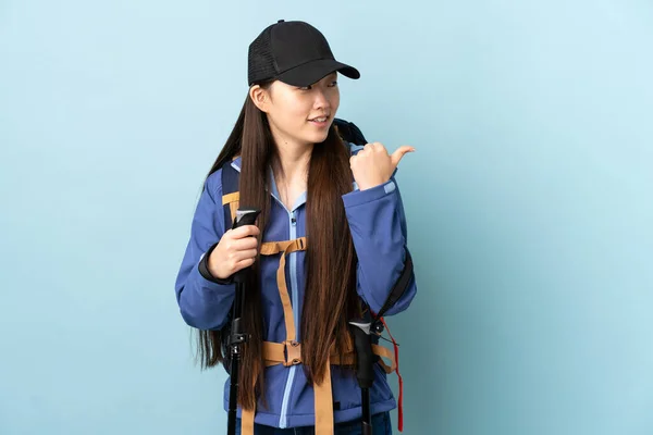 一个年轻的中国姑娘 背着背包 带着远足的杆子 穿过蓝色的孤立的背景 指向侧面 展示自己的产品 — 图库照片
