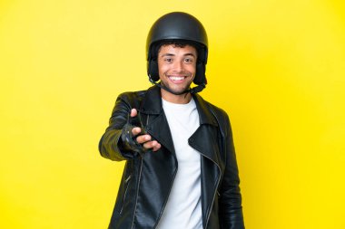 Motosiklet kasklı genç Brezilyalı adam sarı arka planda izole edilmiş, iyi bir anlaşma için el sıkışıyor.