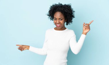 Mavi arka planda izole edilmiş genç Afrikalı Amerikalı kadın parmak uçlarını işaret ediyor ve mutlu