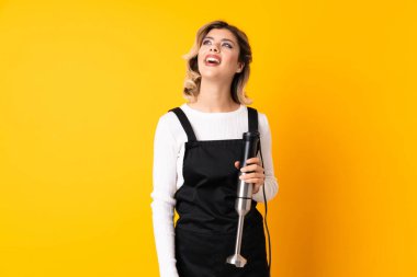 Sarı arka planda izole edilmiş el blenderi kullanan kız gülüyor.