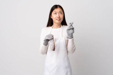 Çinli balıkçı önlük takıyor ve beyaz arka planda çiğ balık tutuyor. Parmaklarını çapraz yapıp en iyisini diliyor.