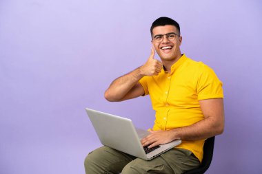 Genç adam dizüstü bilgisayarlı bir sandalyede oturuyor ve baş parmak hareketi yapıyor.