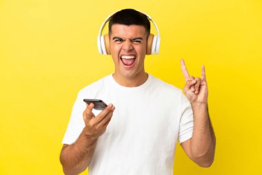 Genç yakışıklı adam, izole edilmiş sarı arka planda gezici bir rock hareketi yaparak müzik dinliyor.