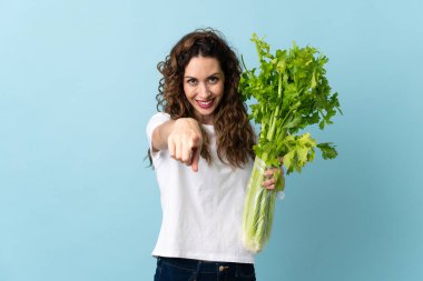 Mavi arka planda izole edilmiş bir kereviz tutan genç kadın kendine güvenen bir ifadeyle seni işaret ediyor.